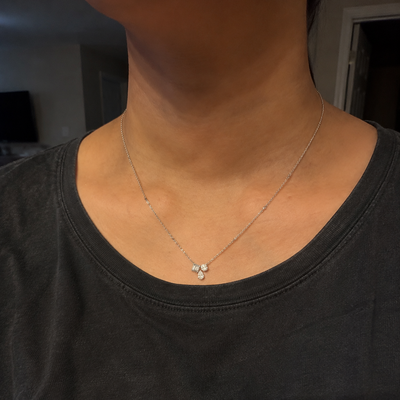 A close-up of a person's neck and upper chest wearing a delicate silver chain necklace with a small three-stone pendant, paired with a dark gray t-shirt. Diamondtine Testimonial